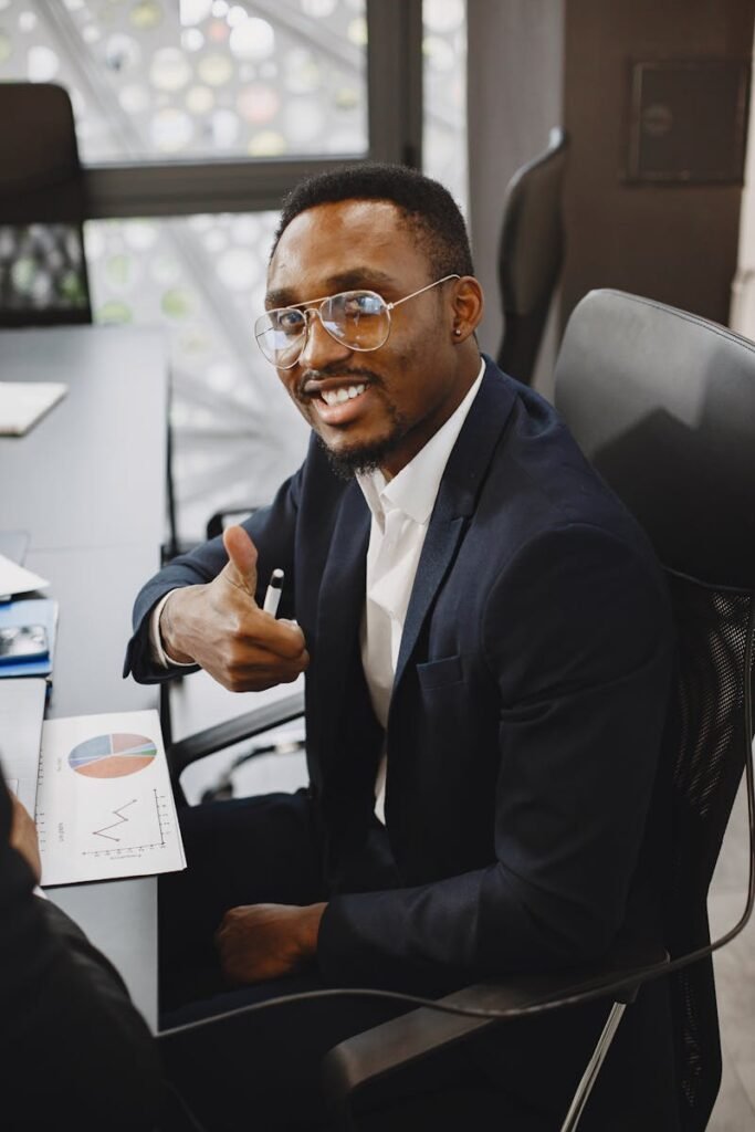 Smiling African American businessman in black suit gives thumbs up in office setting.