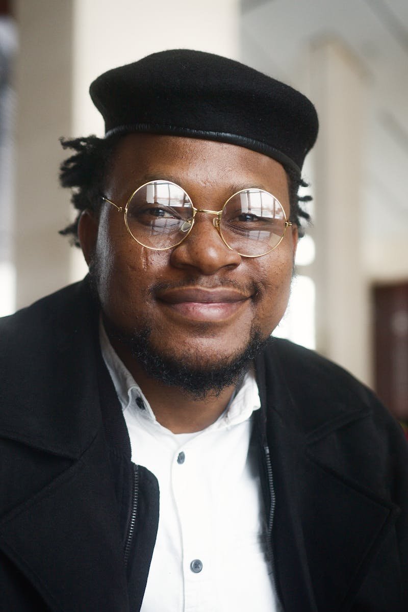 Confident man in glasses with a stylish hat smiling indoors.